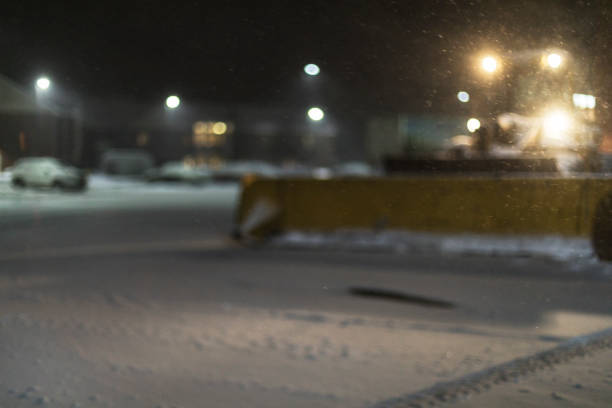 Abstract, intentionally defocused view of a large construction equipment front loader snowplow with a massive snow pusher mounted in front. The loader is about to begin pushing and plowing rapidly gathering and drifting January blizzard snowstorm snow out of parking areas and away from parked vehicles in a large suburban supermarket parking lot near Rochester, New York.