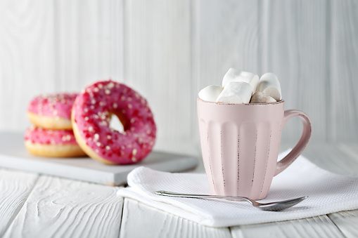 Donuts with pink glaze and colored caramel powder and cocoa with marshmallows on a white wooden background.