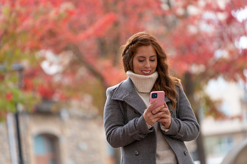 Elegant young woman texting with her phone on the street in autumn with colorful trees on the background