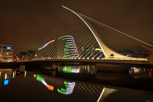 Dublin, Ireland - November 13, 2021: Beautiful wide angle evening view of Samuel Beckett Bridge and The Convention Centre Dublin with ring beam lighting imitating Irish flag and reflection in water