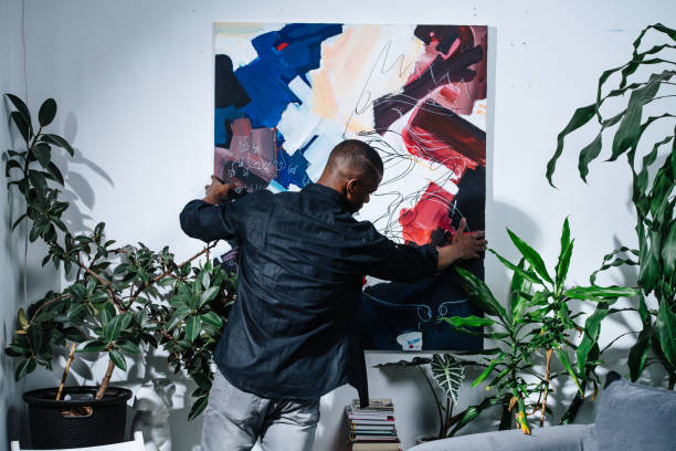 African american painter hanging his abstract colorful piece on the wall. African american painter hanging his abstract colorful piece on the wall. Potted plants all around him. hanging art on wall stock pictures, royalty-free photos & images