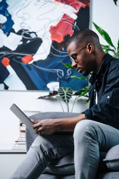 istock Side view african american man sitting on a sofa with a laptop 1358545561