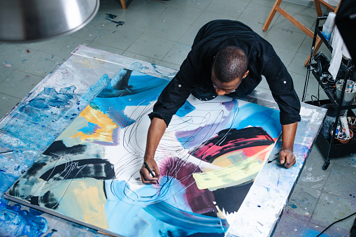 Busy black man drawing lines on a big canvas with painting on a table, leaning over it. He is holding a felt-tip pen. Inside of an apartment.