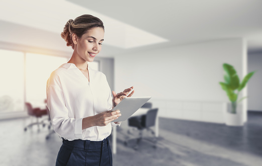 Portrait of smiling young businesswoman using tablet computer in blurry office. Concept of technology and internet. Toned image