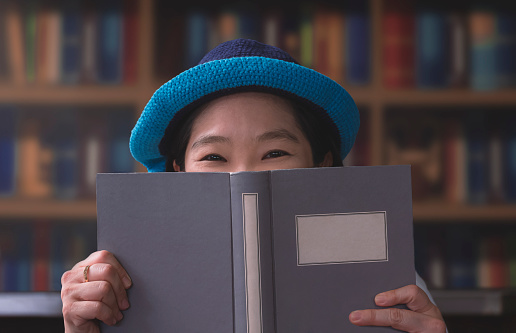 Portrait of Asian woman smiling and looking at camera while hiding her face behind a book on blurred bookshelf background in living room
