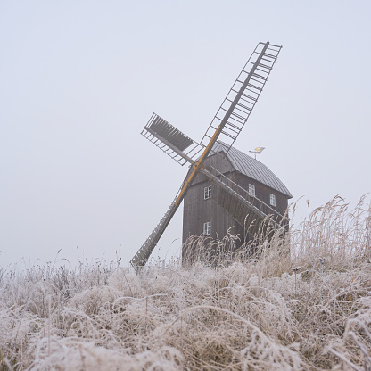 This picturesque wooden windmill has four blades. Located on a hill, it is visible from afar. The tall grass is covered with hoarfrost.