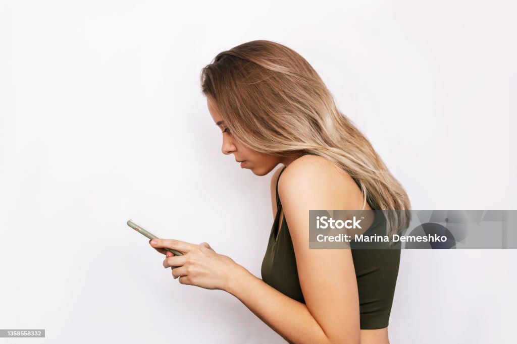 A young woman with a hunched back looks at the mobile phone screen. Incorrect back position A young blonde woman with a hunched back looks at the mobile phone screen isolated on a white background. Incorrect back position, curvature of posture, spine. Dependence on gadgets Back Stock Photo