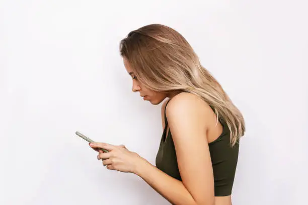 A young blonde woman with a hunched back looks at the mobile phone screen isolated on a white background. Incorrect back position, curvature of posture, spine. Dependence on gadgets