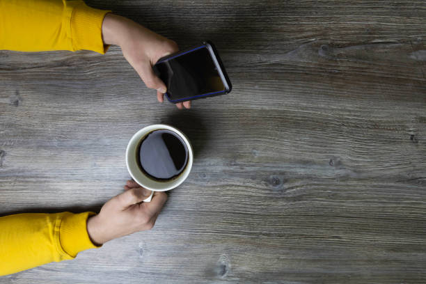 A man drinks coffee in the morning and works on the phone. Men's hands hold a white cup of coffee and a mobile phone in a yellow jacket. The hands lie on a gray wooden surface that is used as a background.
