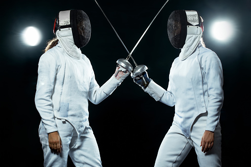 Portrait of Young woman fencer wearing mask and white fencing costume and holding the sword in front of her. Isolated on Black Background.