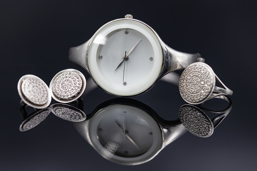 Elegant women's watches in a metal case and jewelry made of silver lie on a black reflective surface