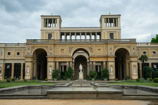 Grand palace in Sanssouci Park, Potsdam, Germany featuring ornate rococo architecture, statues, domed pavilions, and expansive green lawns. Visitors cycle along pathways beneath a vivid blue sky.