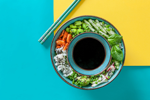 Pan Asian trendy Homemade Vegan Poke Bowl with Rice, edamame, fresh vegetables and soy sauce, served with chopsticks on vivid yellow and turquoise background