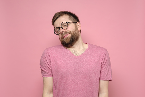 Cute, shy man with glasses hunched shoulders and looked with narrowed eyes at the camera. Studio, pink background.