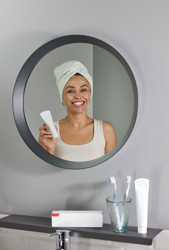 Mid-aged woman's reflection on a bathroom mirror while she is holding a toothpaste