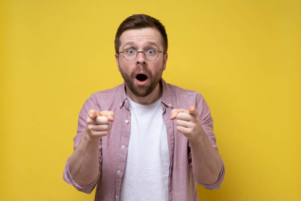 istock Surprised man with glasses points with index fingers and looks with large eyes, with an open mouth. Yellow background. 1358609053
