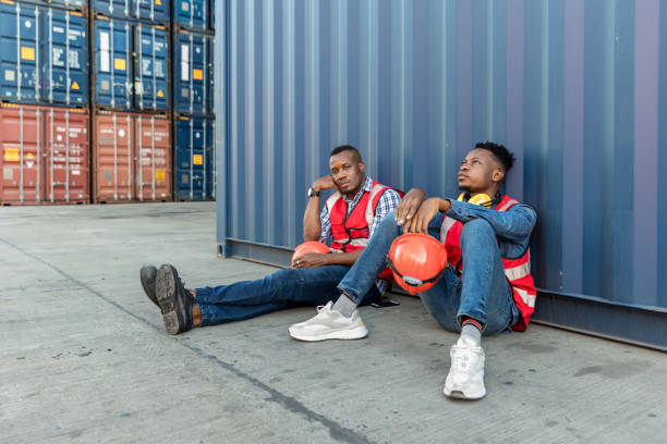 istock American African Technician worker men sit close to cargo container and he look tired and sleepy or lost job. Factory worker man sit close to the cargo container and he look tire and lost job. 1358614323