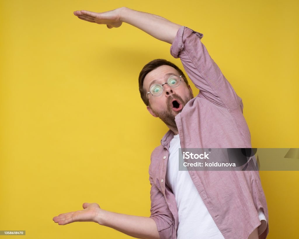 Surprised man in glasses shows a large size with hands, he exaggerates and looks at the camera in amazement. Copy space. Surprised man in glasses shows a large size with hands, he exaggerates and looks at the camera in amazement. Copy space. Isolated, yellow background. Scale Stock Photo