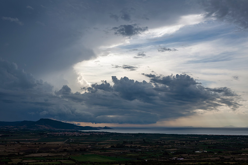 Dark storm supercell clouds above a Mediterranean landscape inland, in the distance visible the sea.