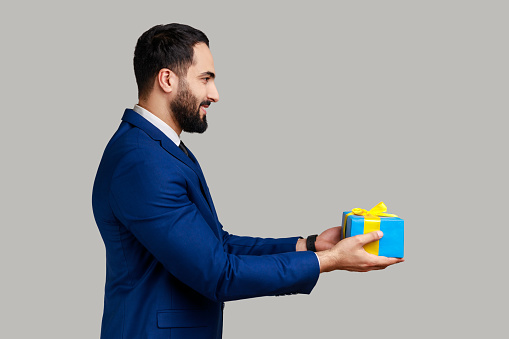 Side view portrait of smiling positive man giving blue wrapped present box, congratulating with holidays, holding gift, wearing official style suit. Indoor studio shot isolated on gray background.