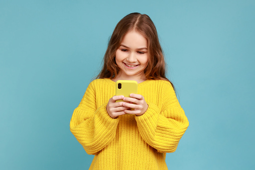 Little girl reading message on smartphone and smiling, satisfied with children mobile application, wearing yellow casual style sweater. Indoor studio shot isolated on blue background.