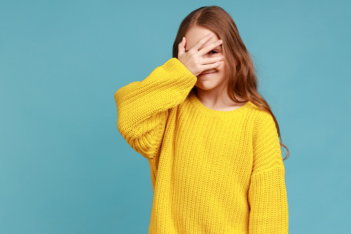 Little girl covers face with hand, looking through fingers, curious and shy about watching secret, wearing yellow casual style sweater. Indoor studio shot isolated on blue background.