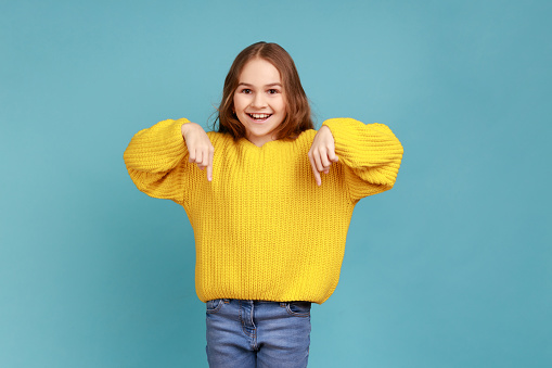 Portrait of cute little girl pointing fingers down, showing place below for commercial information, wearing yellow casual style sweater. Indoor studio shot isolated on blue background.