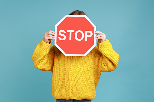 Portrait of unknown little kid covers face with Stop symbol, anonymous child holds red traffic sign, wearing yellow casual style sweater. Indoor studio shot isolated on blue background.