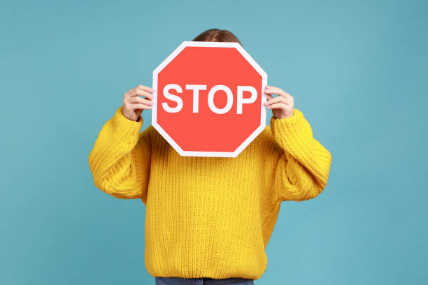 Portrait of unknown little kid covers face with Stop symbol, anonymous child holds red traffic sign. Portrait of unknown little kid covers face with Stop symbol, anonymous child holds red traffic sign, wearing yellow casual style sweater. Indoor studio shot isolated on blue background. stop stock pictures, royalty-free photos & images