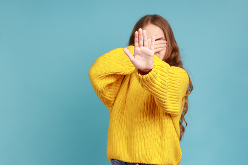 Little girl covers eyes with hand, shows stop gesture,feels shame or fear to watch forbidden content, wearing yellow casual style sweater. Indoor studio shot isolated on blue background.