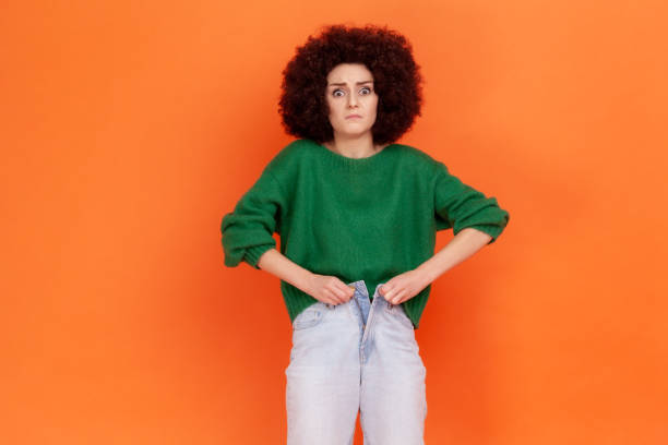 ritratto di giovane donna adulta triste sconvolta con acconciatura afro che indossa un maglione verde stile casual incapace di abbottonare i jeans a causa dell'aumento di peso. - belly button foto e immagini stock