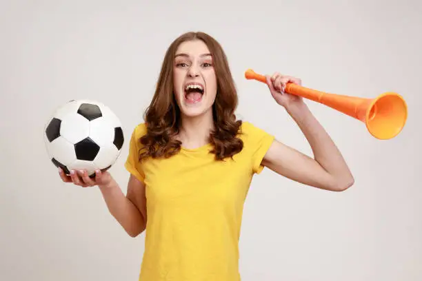 Photo of Portrait of cheerful woman of young age with brown hair in yellow t-shirt holding big bullhorn and football ball, celebrating championship beginning.