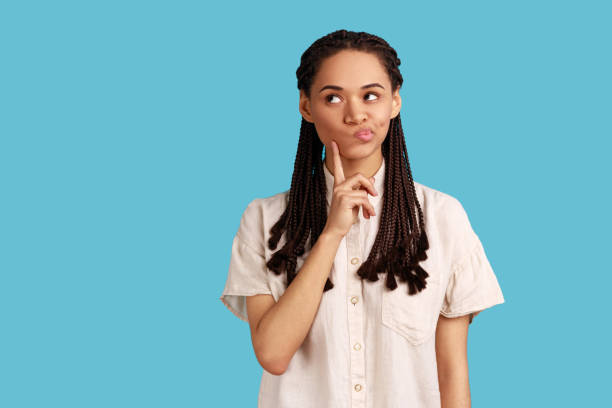 Woman with black dreadlocks keeps index finger on cheek, considers something, has pensive expression Portrait of thoughtful woman with black dreadlocks keeps index finger on cheek, considers something, has pensive expression, wearing white shirt. Indoor studio shot isolated on blue background. asking stock pictures, royalty-free photos & images