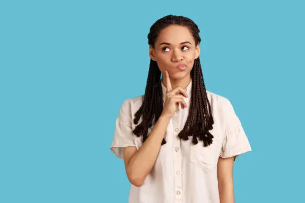 Portrait of thoughtful woman with black dreadlocks keeps index finger on cheek, considers something, has pensive expression, wearing white shirt. Indoor studio shot isolated on blue background.