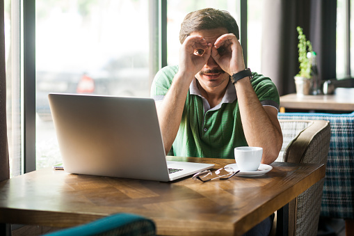 Portrait of businessman wearing green T-shirt, working online on laptop, looking through fingers shaped like binoculars and expressing positive. Indoor shot near big window, cafe background.