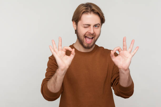 Man showing ok gesture with fingers, approving work, satisfied with quality, looks winking at camera Portrait of man with beard wearing sweatshirt, showing ok gesture with fingers, approving work, satisfied with quality, looking winking at camera. Indoor studio shot isolated on gray background. young man wink stock pictures, royalty-free photos & images