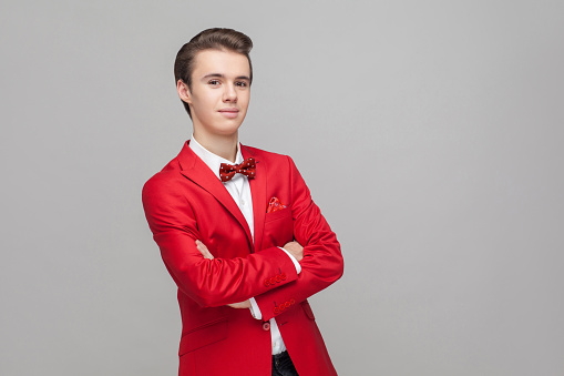 Portrait of trendy handsome gentleman with stylish hairdo wearing red tuxedo and bow tie standing with crossed hands and positive calm expression. indoor studio shot isolated on gray background