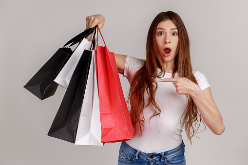 Shocked amazed dark haired woman pointing finger at paper bags in her hand, surprised with shopping, low prices good quality, wearing white T-shirt. Indoor studio shot isolated on gray background.