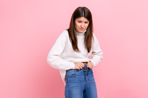 Portrait of unhappy sad young adult woman gaining weight, cant wearing her jeans, being overweight, wearing white casual style sweater. Indoor studio shot isolated on pink background.