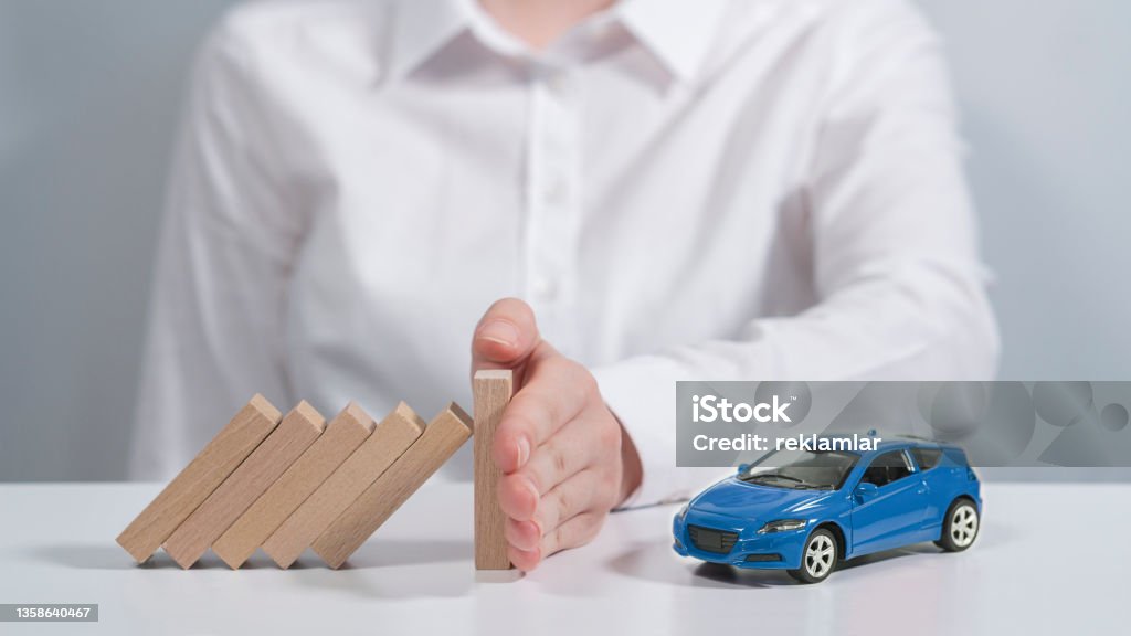 Next to the little blue car is a person's hand that prevents dangers from coming to her. Authoritative person sitting in front of gray background. His face is showing. Insuring the vehicle safely. Car insurance protects any damage to the vehicle. The hand of the person wearing the white shirt prevents the boards from damaging the falling vehicle. Car Insurance Stock Photo
