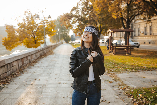 Beautiful happy young woman in a leather jacket and with a bandana on her forehead enjoys a sunny autumn day in public park