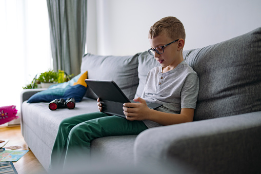 Smiling boy watching cartoon on digital tablet while sitting on a couch in a living room.