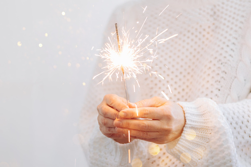 A girl in a white sweater holds a burning sparkler. Christmas New Year party holiday concept. Selective focus, light background, copy space