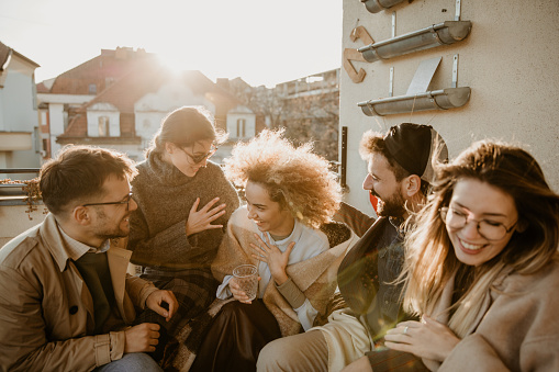 Image of a small group of five, having big laughter while sitting on the small balcony of their working space, their brainstorming spot and enjoying the last days of the year, on a cold and sunny winter day. They’re all looking close and warm, stylishly dressed, some wearing sunglasses and some eyeglasses, loving each other’s company