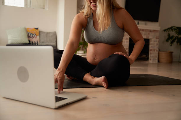 mujer caucásica embarazada sonriendo, sentada en una esterilla de yoga en casa a punto de comenzar el tutorial de acondicionamiento físico en línea - ewan fotografías e imágenes de stock