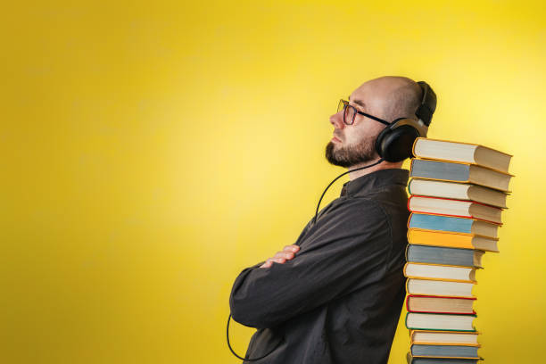 istruzione e tecnologie moderne. il concetto di audiolibri. un uomo con occhiali, camicia e auricolari si appoggiava a una pila di libri. sfondo giallo. spazio di copia - mid adult audio foto e immagini stock