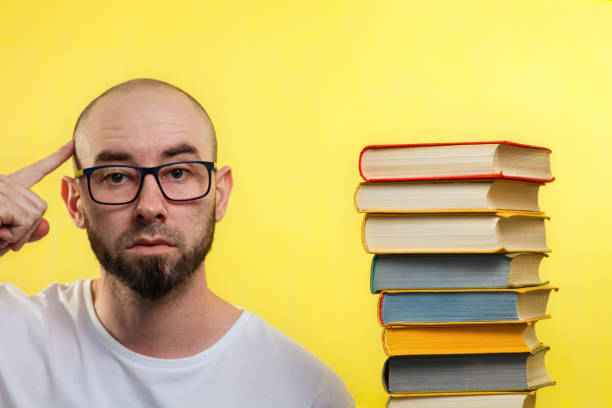 concepto de educación. hombre calvo barbudo con gafas, señalando con el dedo a su cabeza. cerca hay una pila de libros. fondo amarillo - confusion single object human finger one person fotografías e imágenes de stock