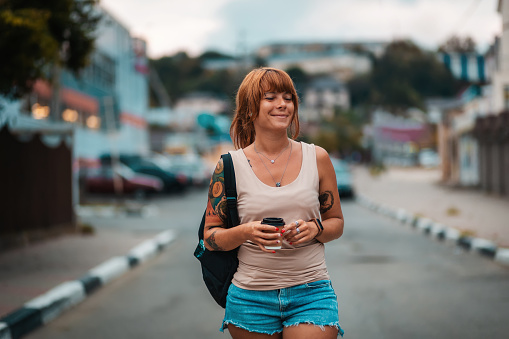 Portrait of happy young woman with tattoo walking with a cup of coffee in her hands. Evening stroll and vacation.