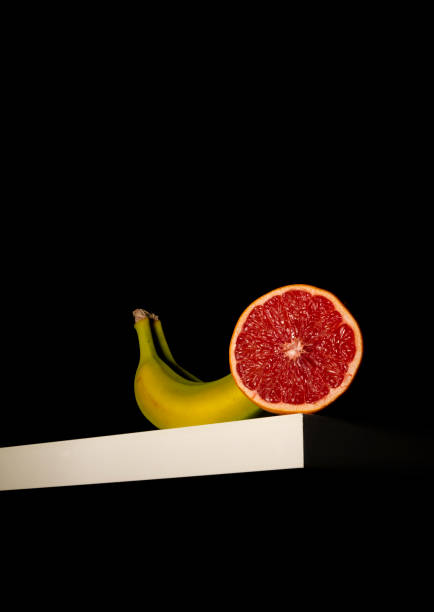 istock Still life of banana and grapefruit on white table against black background 1358717038