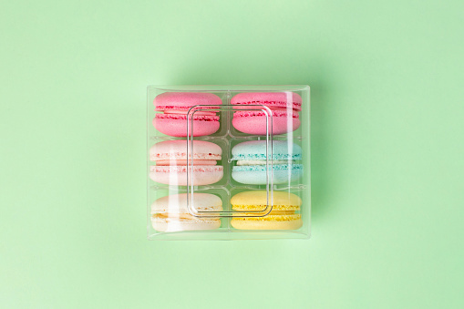 Macaroons of various colors in transparent plastic container on mint green background, studio shot